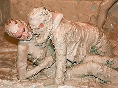 Two women are wrestling each other in a bath filled with mud. They are watched by a referee and another group 