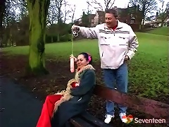A girl is sitting on a bench in the park when a guy comes up from behind her, dangling a rubber dick in front of her f